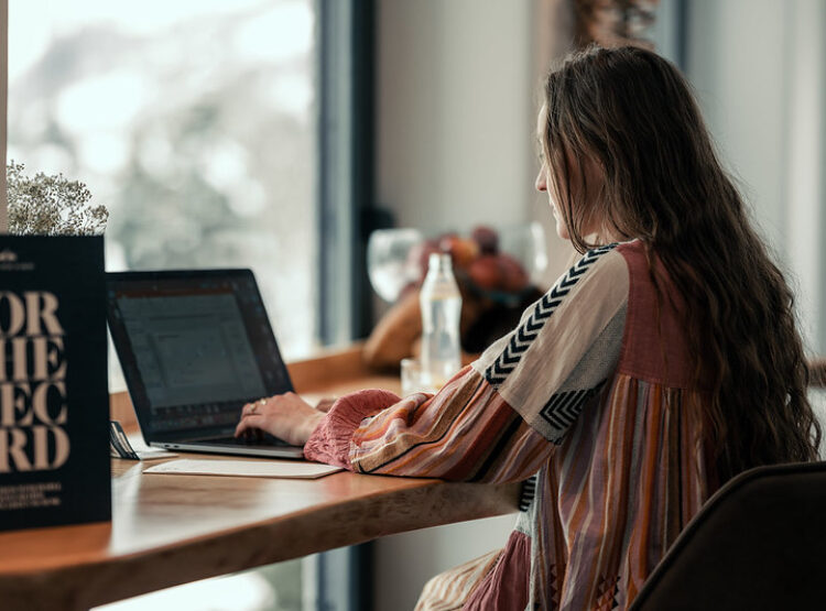 Eine Frau mit langen Haaren arbeitet an einem Laptop an einem Holztisch in der Nähe eines Fensters mit natürlichem Licht.
