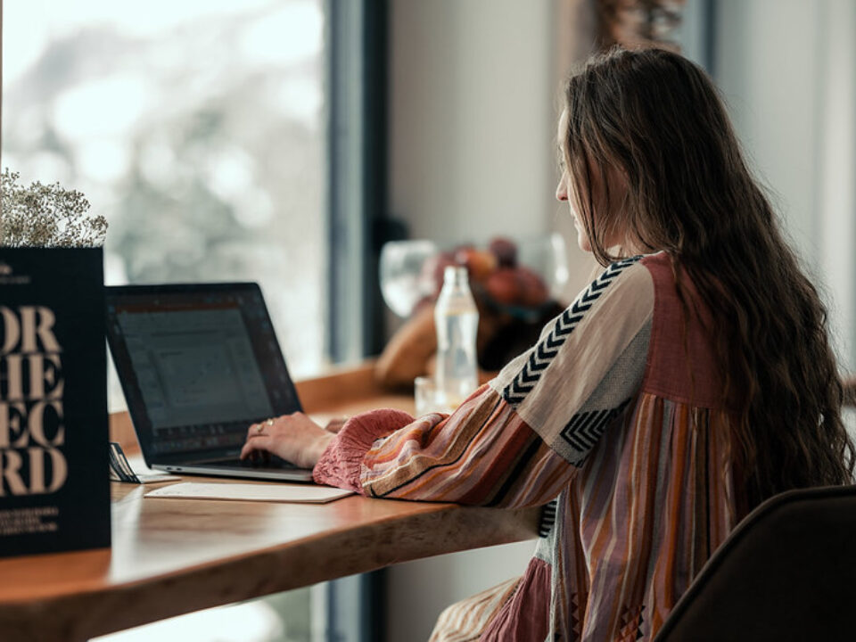 Eine Frau mit langen Haaren arbeitet an einem Laptop an einem Holztisch in der Nähe eines Fensters mit natürlichem Licht.