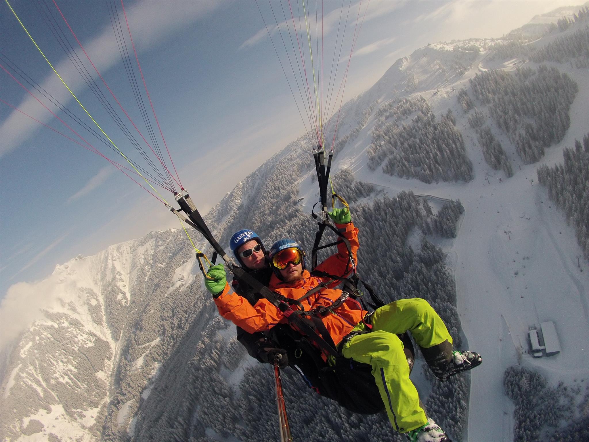 Zwei Personen, die an einem klaren Tag über verschneite Berge und Wälder gleiten, tragen helle Winterkleidung.