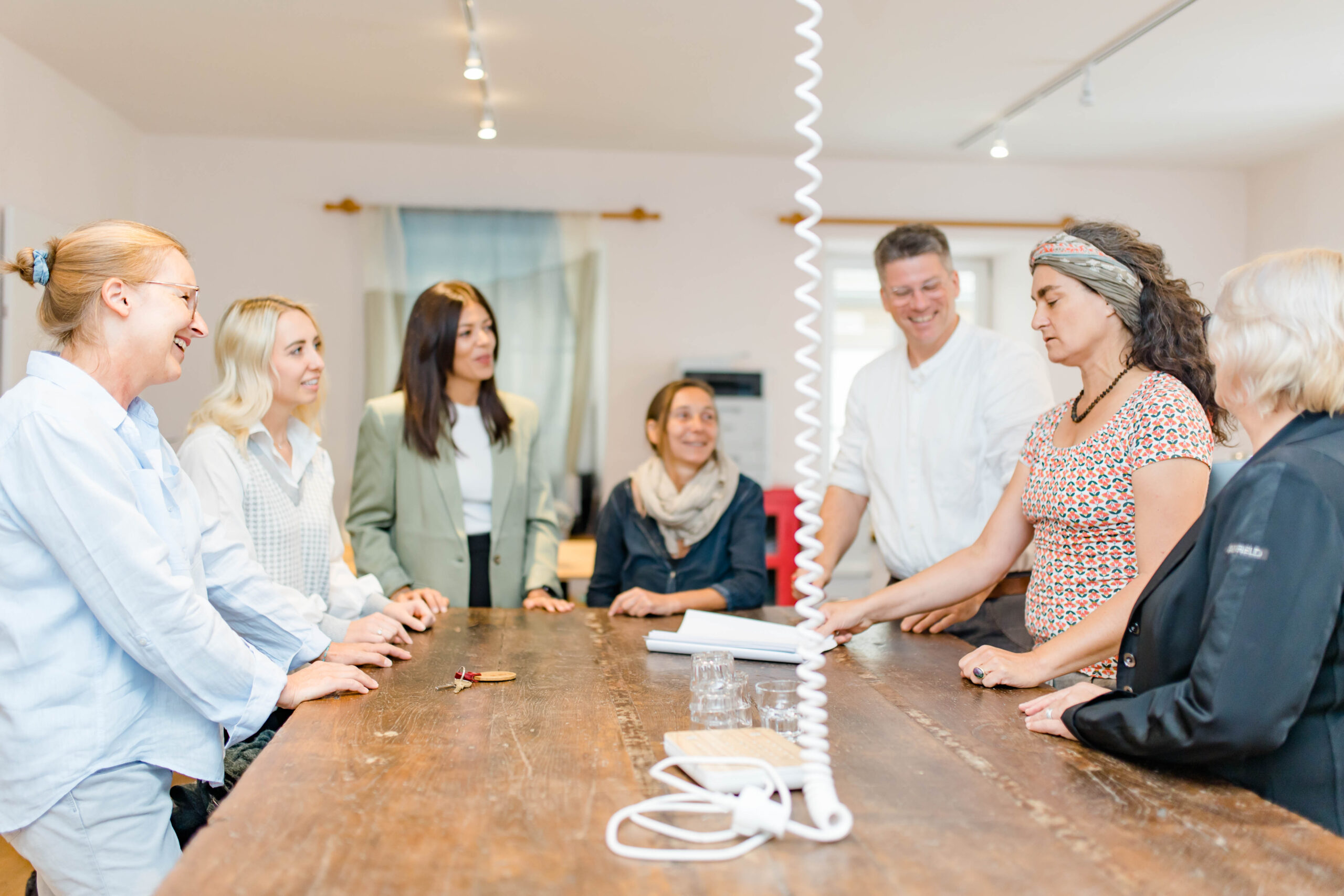 Sechs Personen stehen in einem hellen Raum um einen Holztisch herum und lächeln, als würden sie eine Diskussion führen.