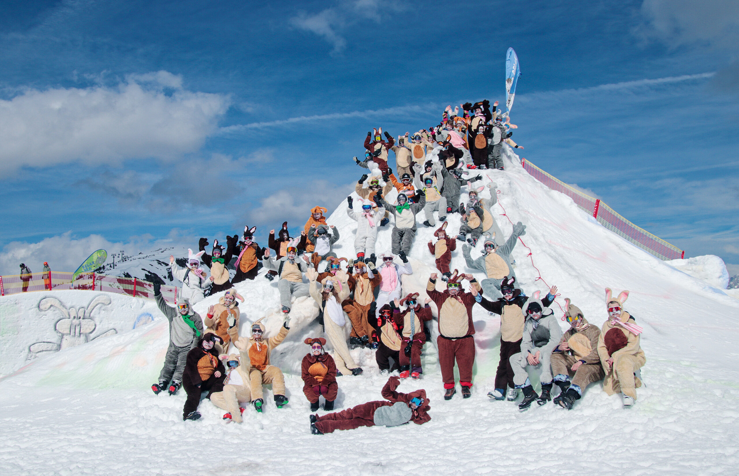 Eine große Gruppe von Menschen in Tierkostümen posiert auf einem verschneiten Hügel unter einem blauen Himmel.