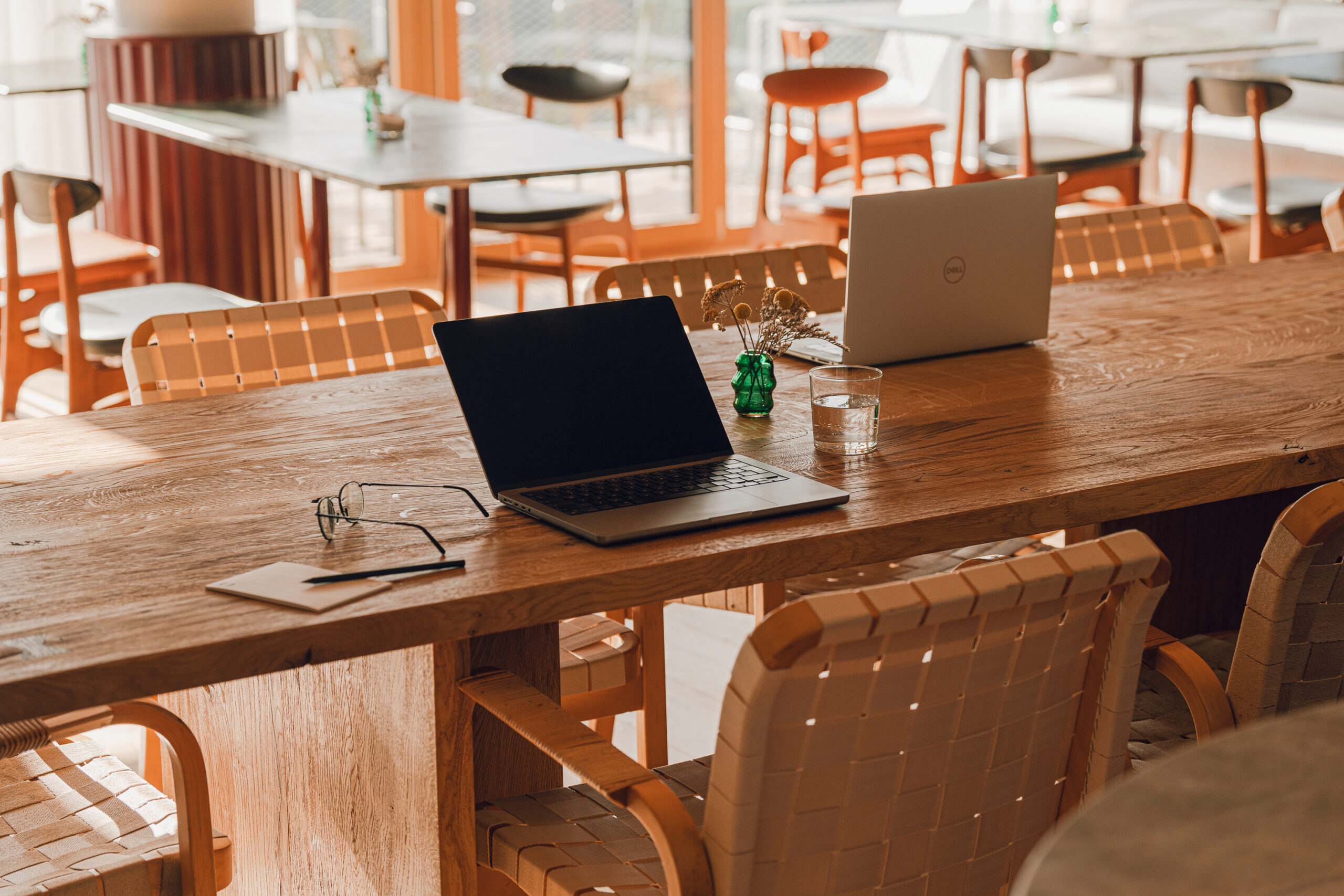 Ein Holztisch mit zwei Laptops, Gläsern, einem Notebook und einem Glas Wasser in einem sonnendurchfluteten Café.