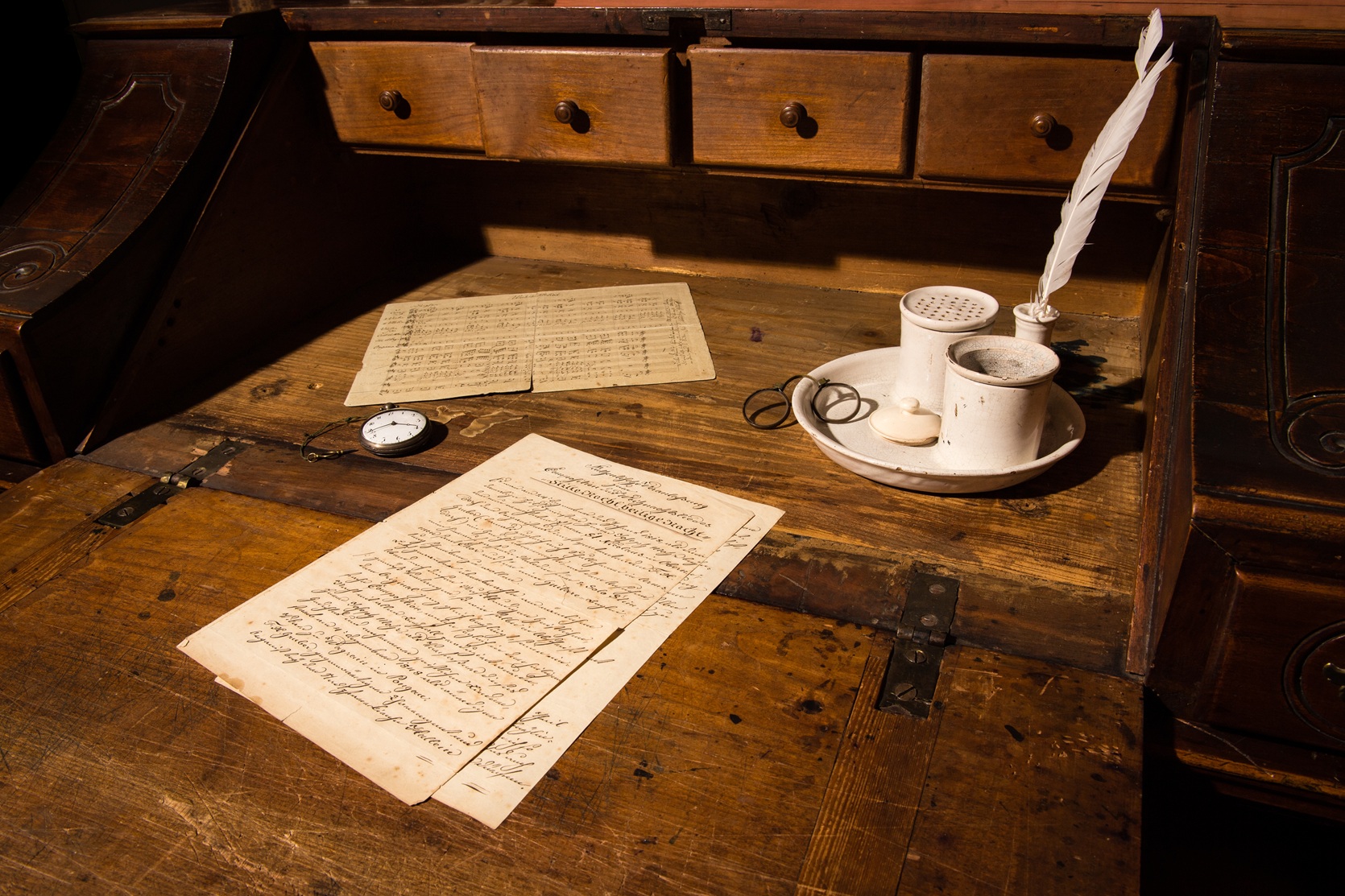 Alter Holzschreibtisch mit handgeschriebenen Briefen, einer Taschenuhr, einer Feder in Tinte und einer Kerze auf einem Tablett.