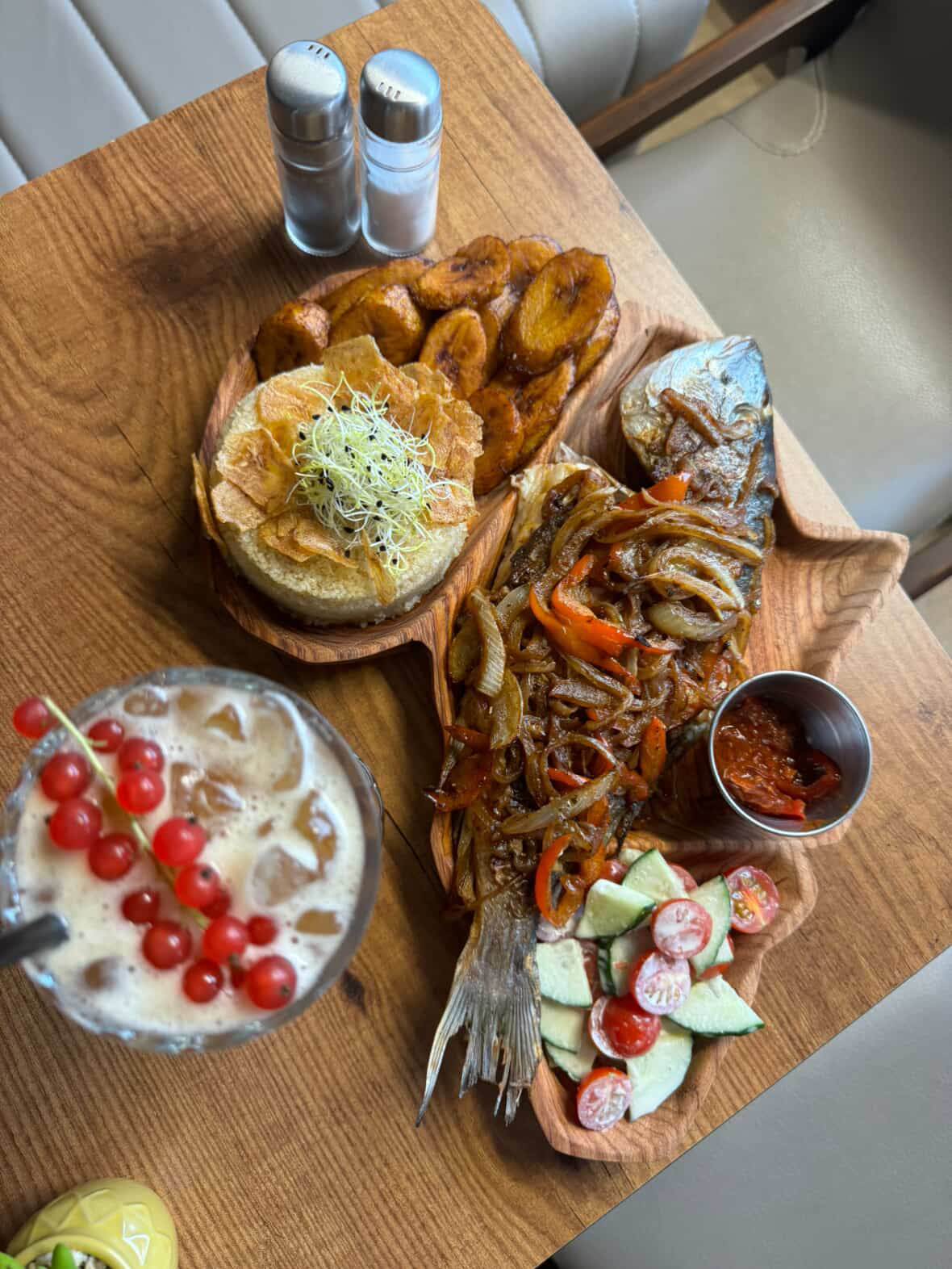 Holztisch mit gegrilltem Fisch, gebratenen Kochbananen, Sandwich, Gurkensalat, Chilisauce und eisgekühltem Getränk mit Beeren im Le Nimba. 