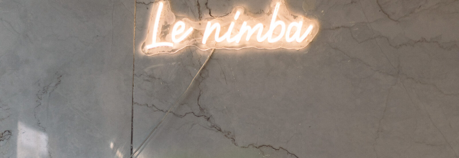 Neonschild mit der Aufschrift Le nimba auf einer grauen Marmorwand, mit einem verschwommenen grünen Objekt am unteren Rand.