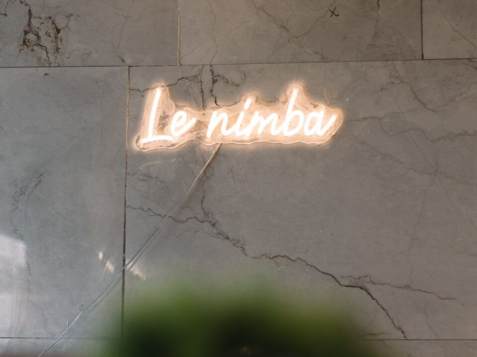 Neonschild mit der Aufschrift Le nimba auf einer grauen Marmorwand, mit einem verschwommenen grünen Objekt am unteren Rand.