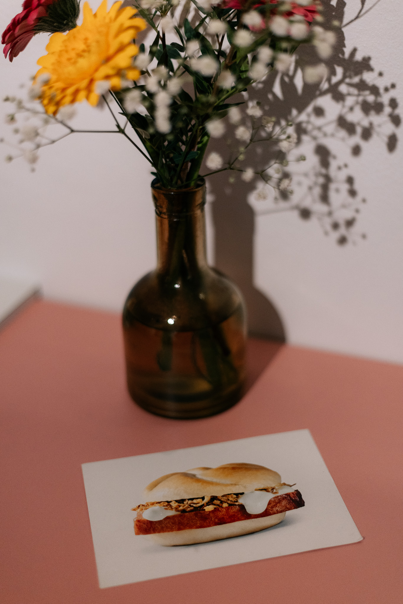 Eine braune Glasvase mit Blumen neben einer Karte, die ein Sandwich auf einer rosa-weißen Fläche zeigt.