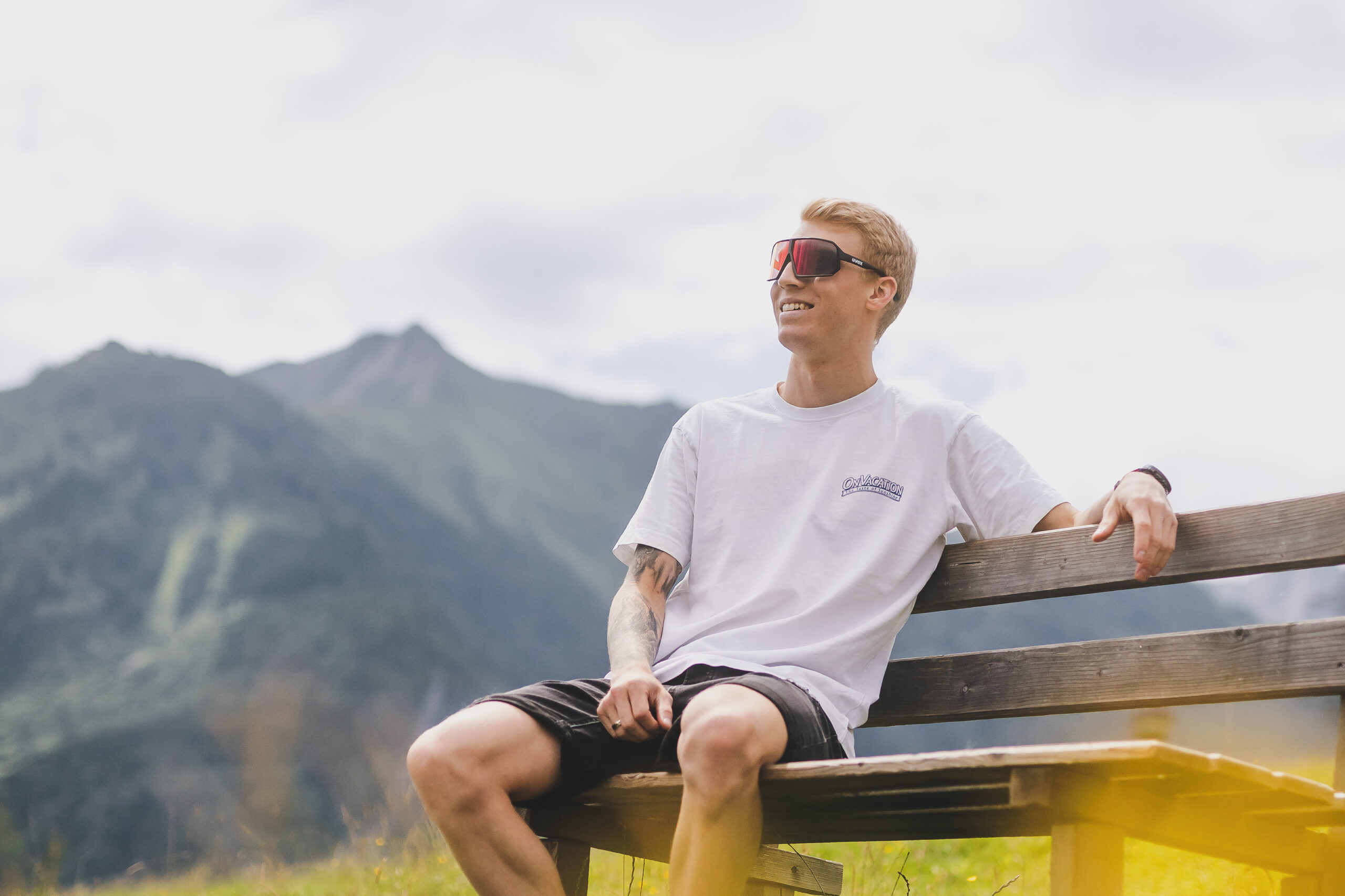 Junger Mann mit Sonnenbrille auf einer Bank sitzend, lächelnd, mit Bergen und bewölktem Himmel im Hintergrund.
