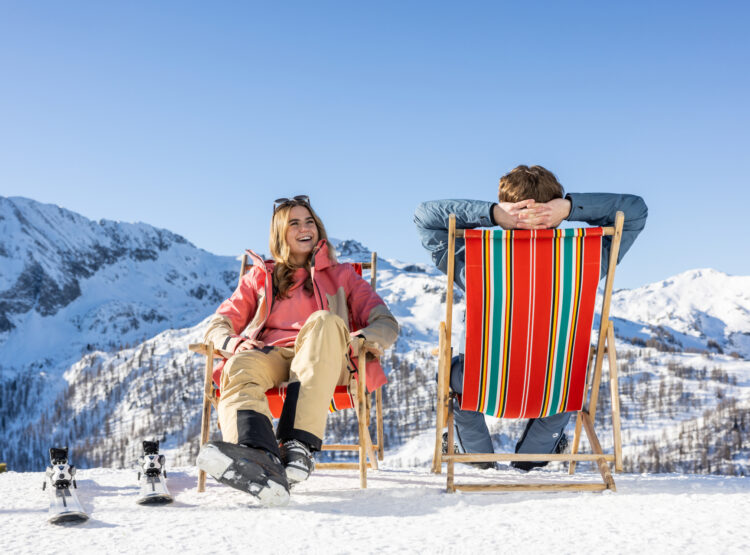 Zwei Personen entspannen sich auf Liegestühlen im Schnee, mit verschneiten Bergen und blauem Himmel im Hintergrund.