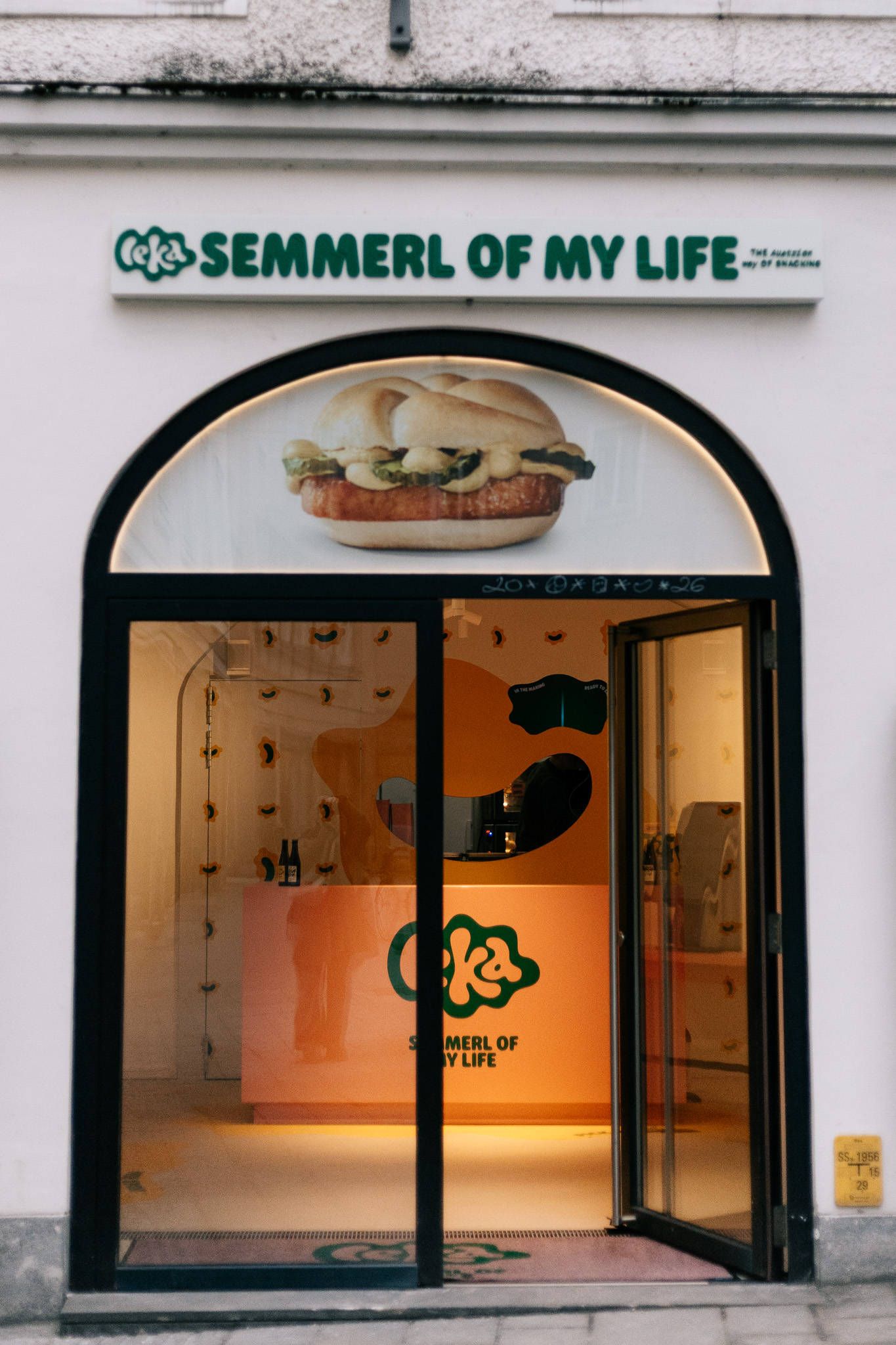 Schaufenster mit "Semmerl of My Life"-Schild, Sandwich-Image und einem pastellfarbenen Innenraum, der durch die offene Tür sichtbar ist.