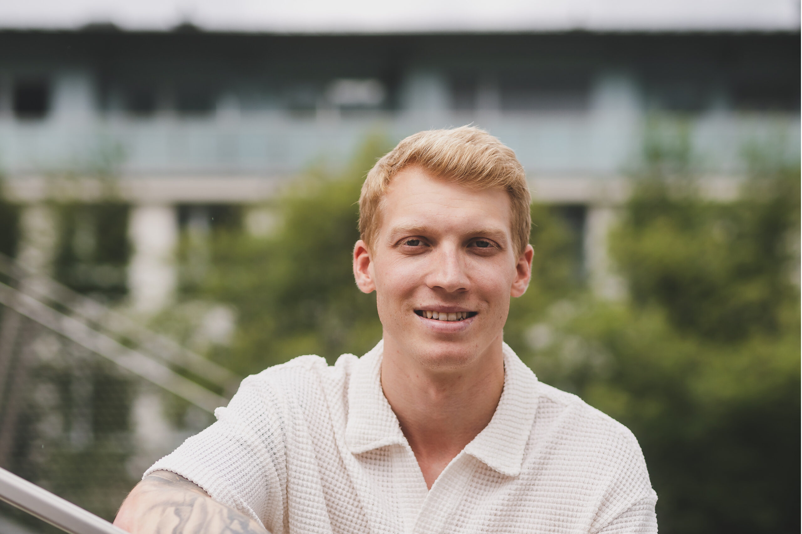 Ein lächelnder junger Mann mit blondem Haar und weißem Hemd steht im Freien, mit Grünflächen und Gebäuden im Hintergrund.