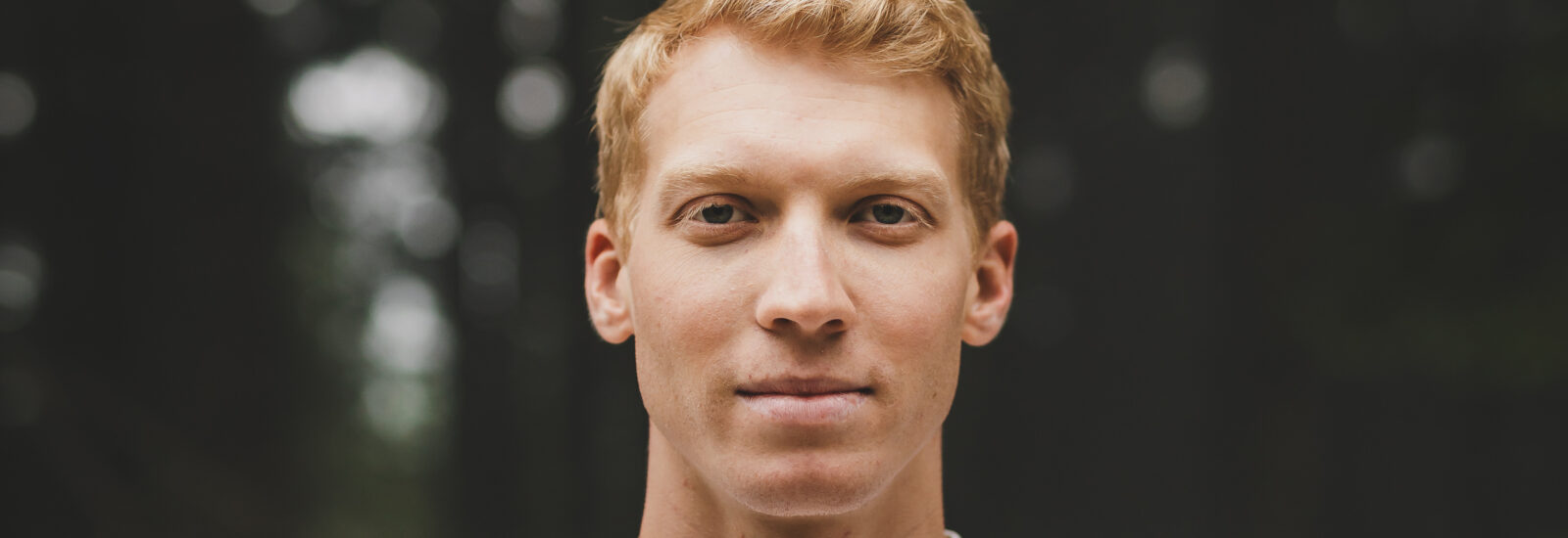 Junger Mann mit kurzen blonden Haaren und weißem Hemd, der im Freien steht, wobei die Bäume im Hintergrund verschwimmen.