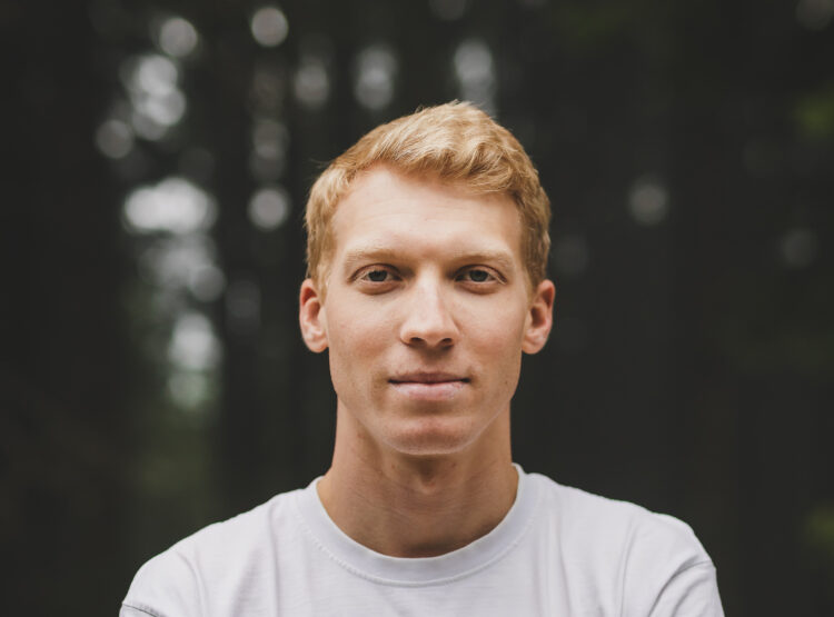 Junger Mann mit kurzen blonden Haaren und weißem Hemd, der im Freien steht, wobei die Bäume im Hintergrund verschwimmen.