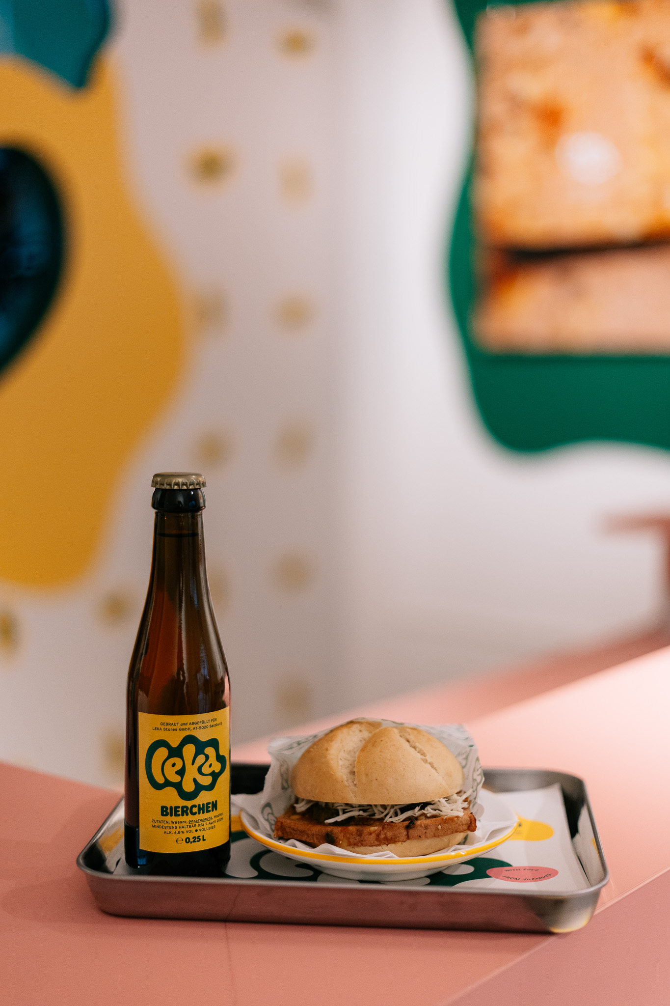 Eine Flasche Ceka Bierchen und ein Sandwich auf einem Tablett auf einem rosafarbenen Tresen mit buntem Dekor im Hintergrund.
