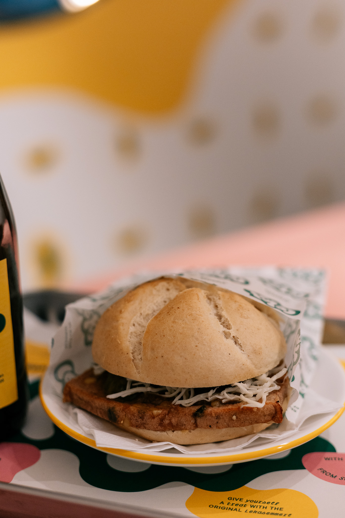 Ein Sandwich mit geriebenem Käse und einer dicken Frikadelle auf einem Teller, daneben eine teilweise sichtbare Flasche.