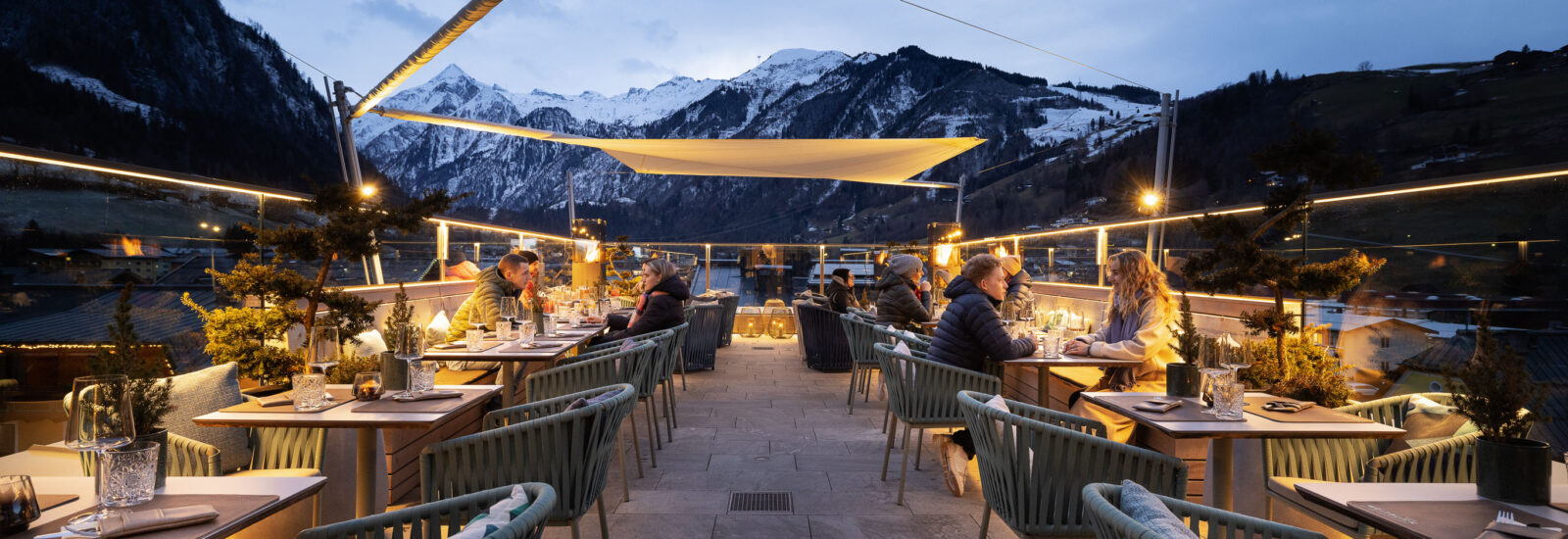 Menschen speisen in der Abenddämmerung auf einer Dachterrasse mit Blick auf die Berge und warmer Beleuchtung.