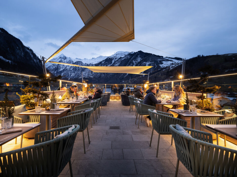 Menschen speisen in der Abenddämmerung auf einer Dachterrasse mit Blick auf die Berge und warmer Beleuchtung.