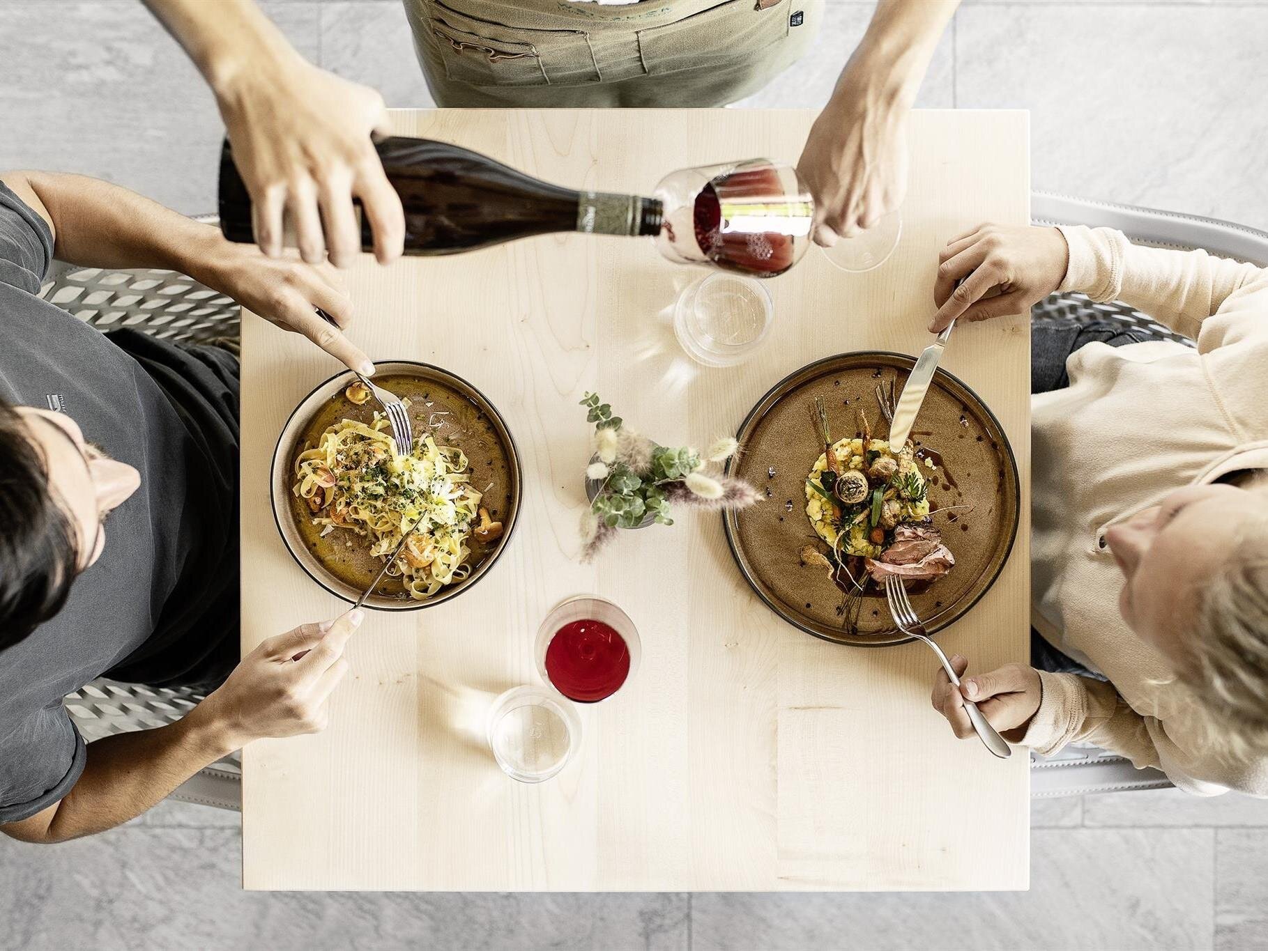 Blick von oben auf zwei Personen, die an einem hellen Holztisch Wein einschenken und Essen auf Tellern servieren.