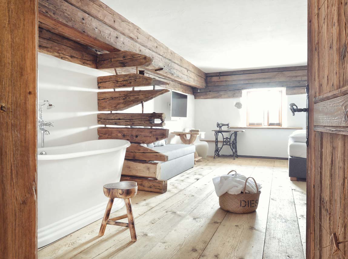 Rustikales Schlafzimmer mit einer freistehenden Badewanne, Holzbalken, einem Hocker und einer Nähmaschine am hellen Fenster.