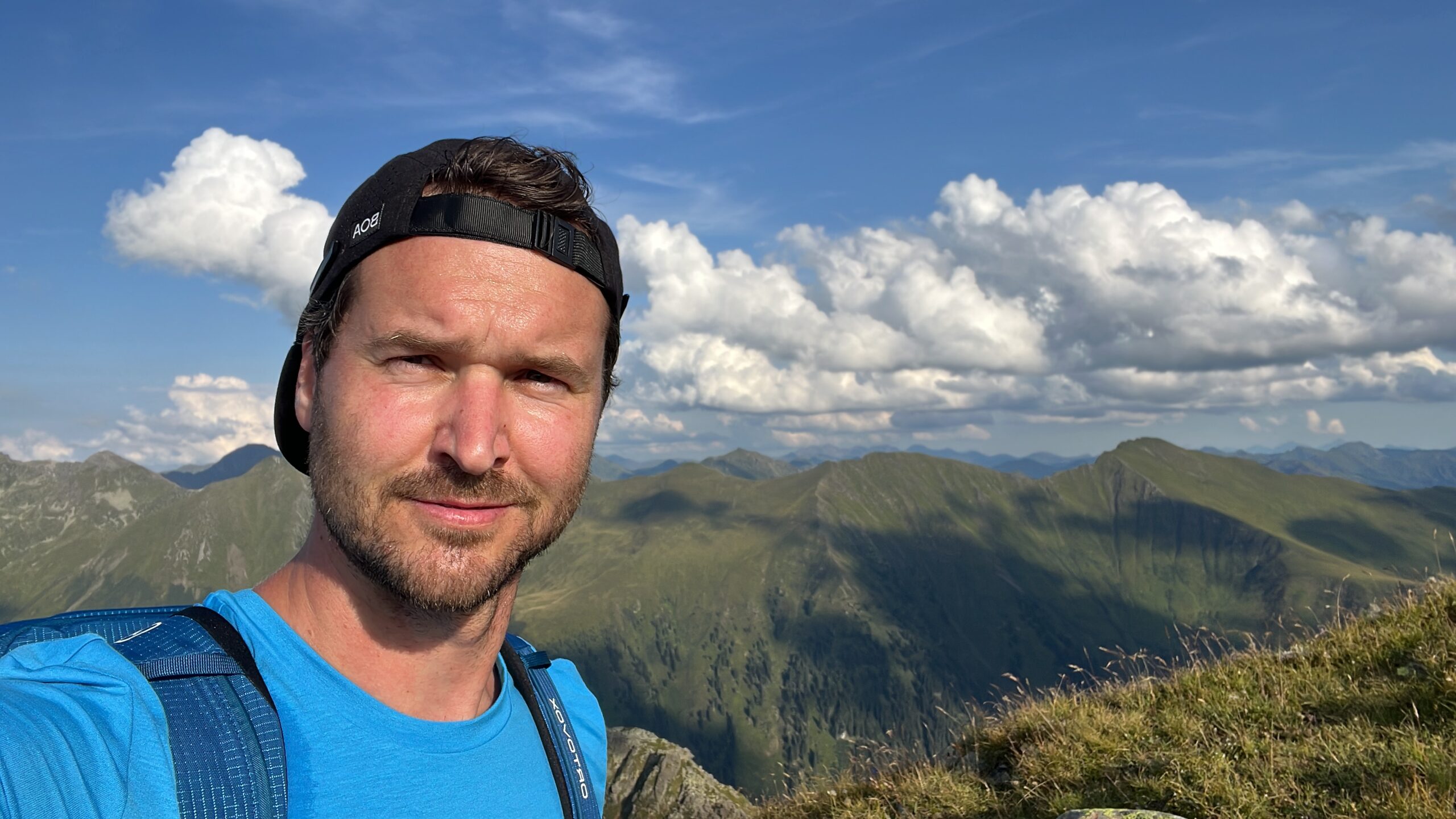 Mann in blauem Hemd und schwarzer Mütze macht ein Selfie mit Bergen und Wolken im Hintergrund.