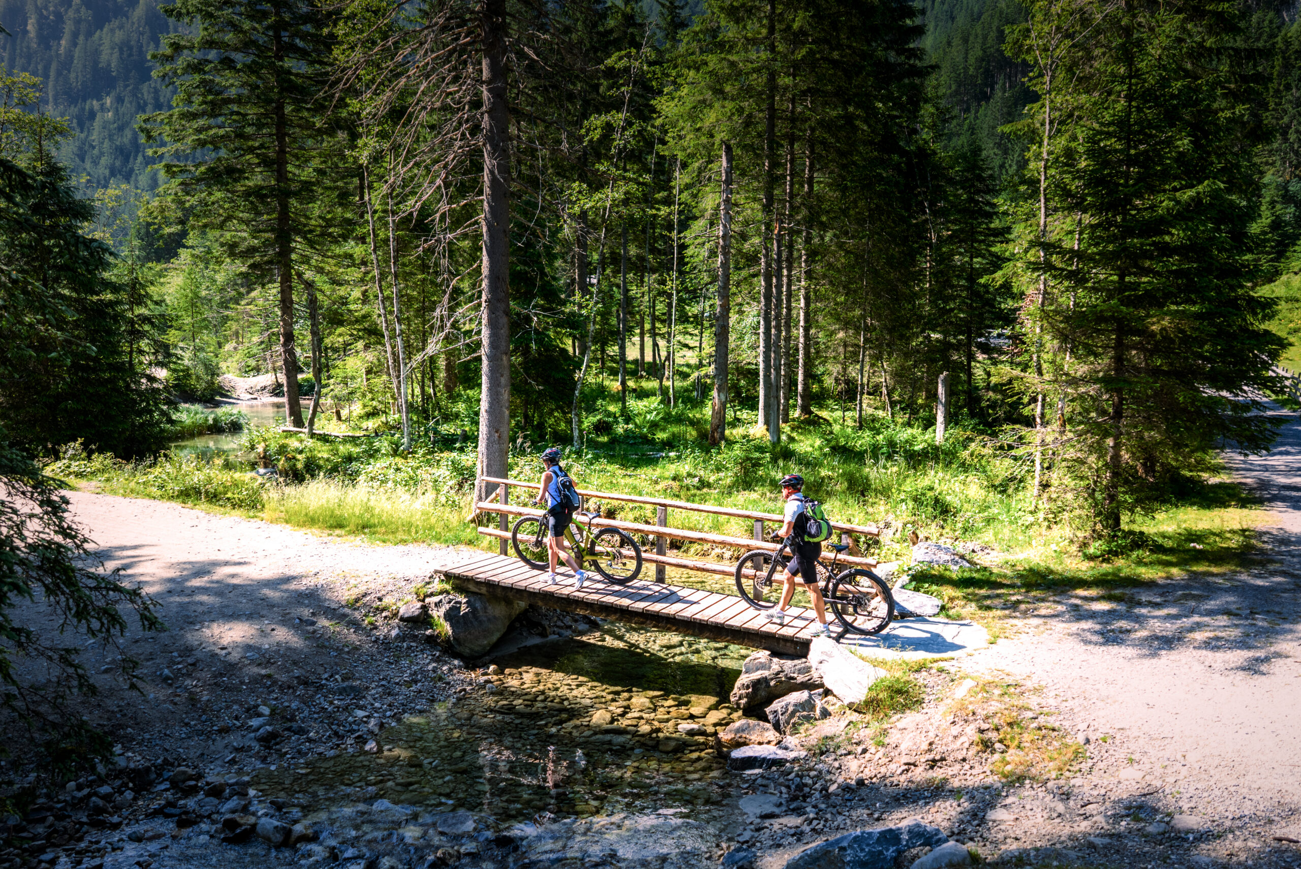 Zwei Radfahrer machen eine Pause auf einer kleinen Holzbrücke in einem sonnenbeschienenen Wald mit einem felsigen Bach unter ihnen.