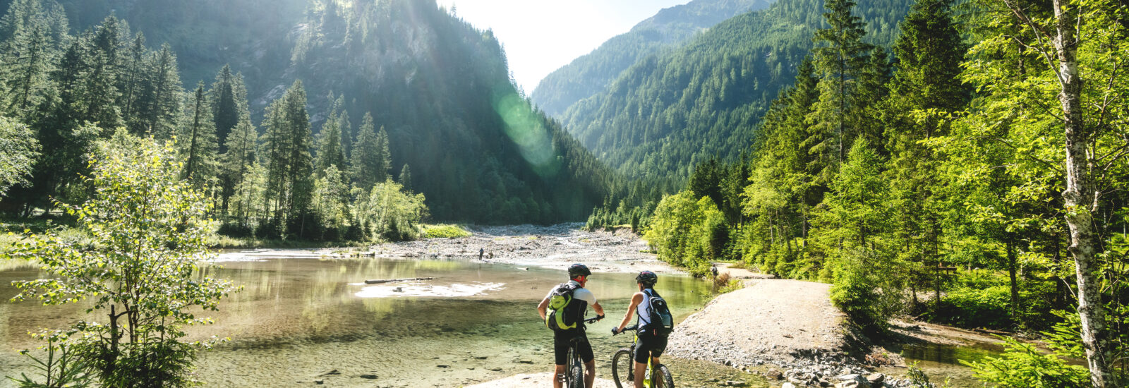Zwei Radfahrer halten an einem klaren Fluss an, der von üppigen grünen Wäldern und Bergen im hellen Sonnenlicht umgeben ist.