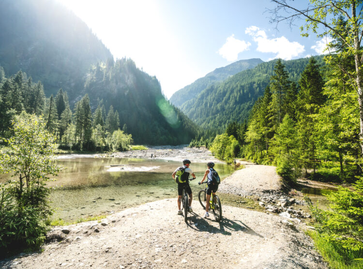 Zwei Radfahrer halten an einem klaren Fluss an, der von üppigen grünen Wäldern und Bergen im hellen Sonnenlicht umgeben ist.