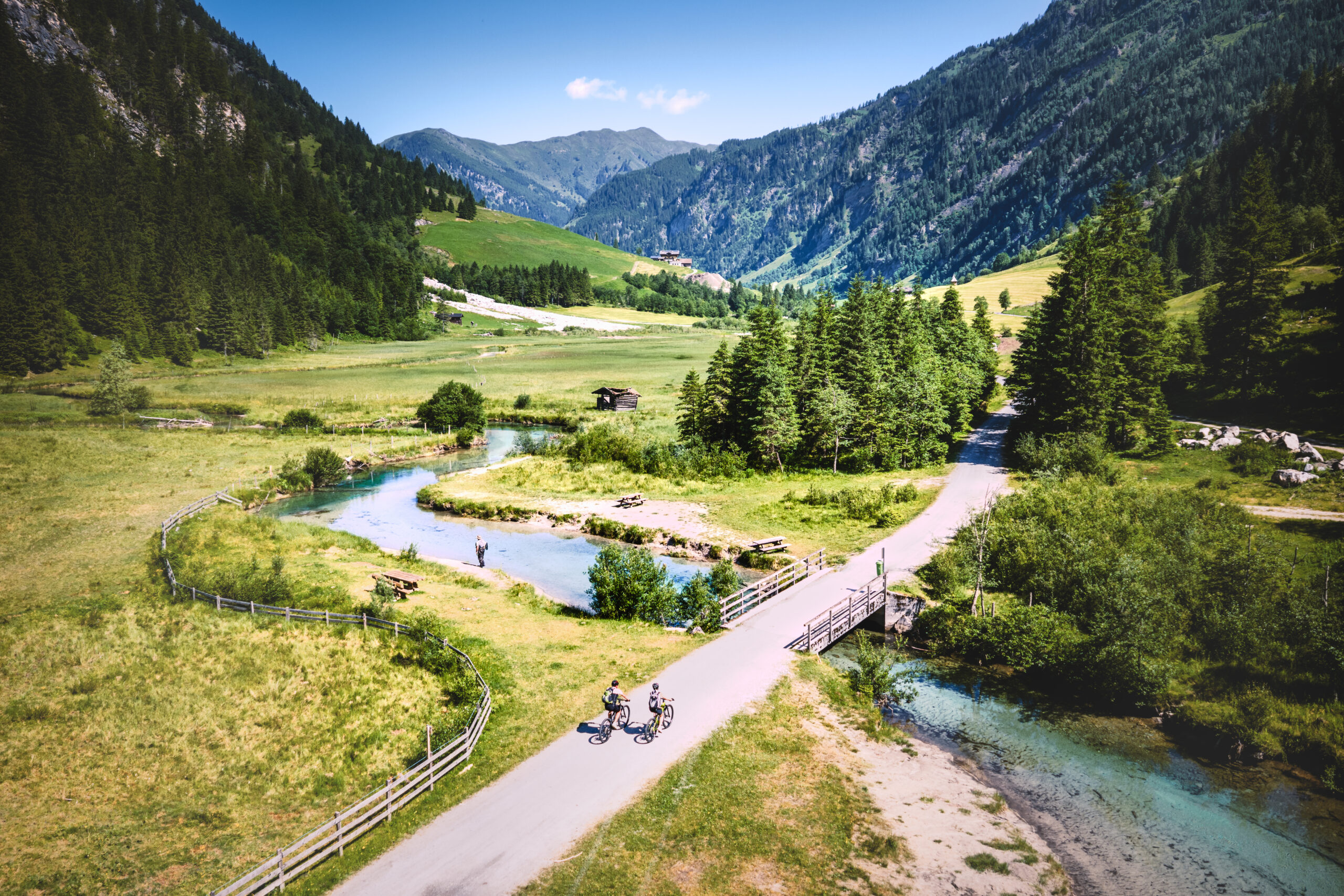 Zwei Radfahrer fahren auf einer Straße durch ein grünes Bergtal mit einem Bach und Bäumen unter einem klaren blauen Himmel.