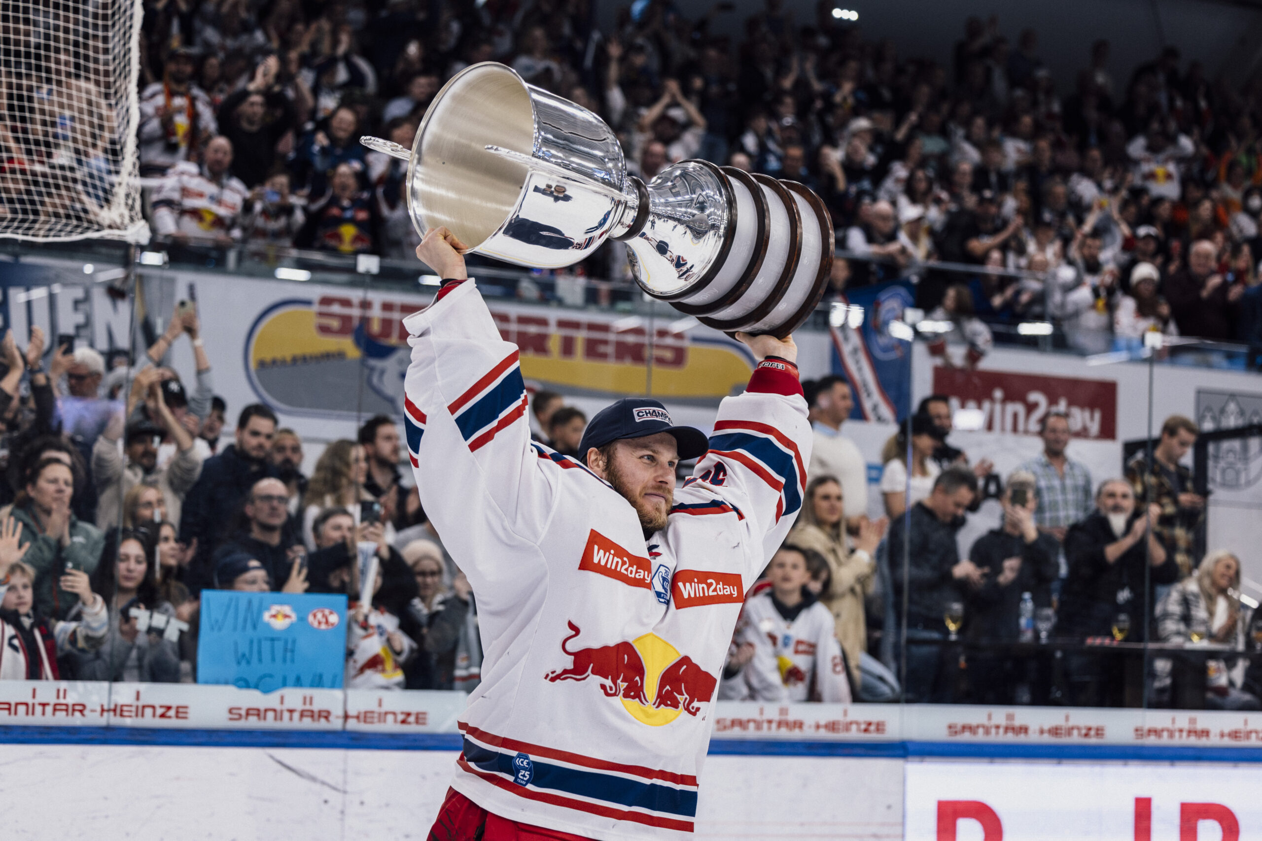 Ein Eishockeyspieler im Red Bull-Trikot hebt eine große Trophäe in die Höhe, während im Hintergrund eine jubelnde Menge jubelt.
