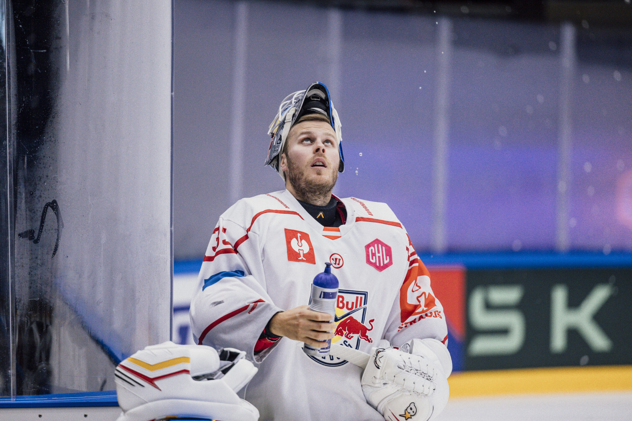Eishockeytorwart in weißer Ausrüstung und mit Helm hält eine Wasserflasche und schaut während eines Spiels nach oben.