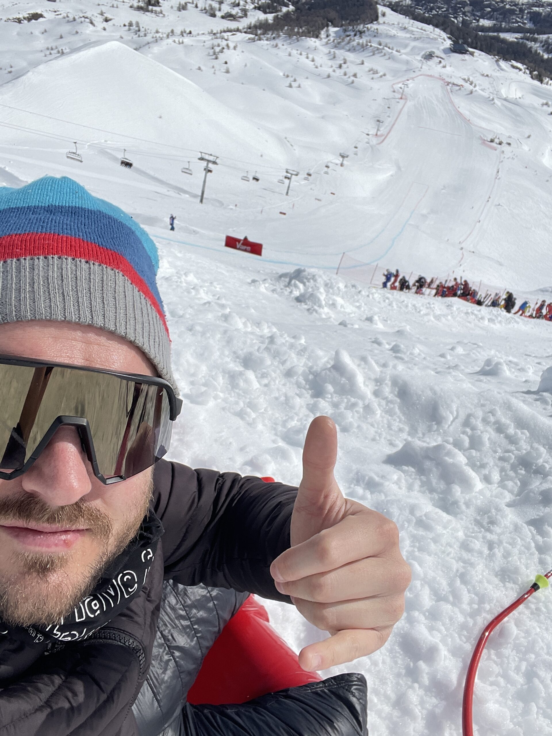 Mann mit Sonnenbrille und Winterausrüstung zeigt Daumen nach oben, während er auf einer verschneiten Piste in einem Skigebiet sitzt.