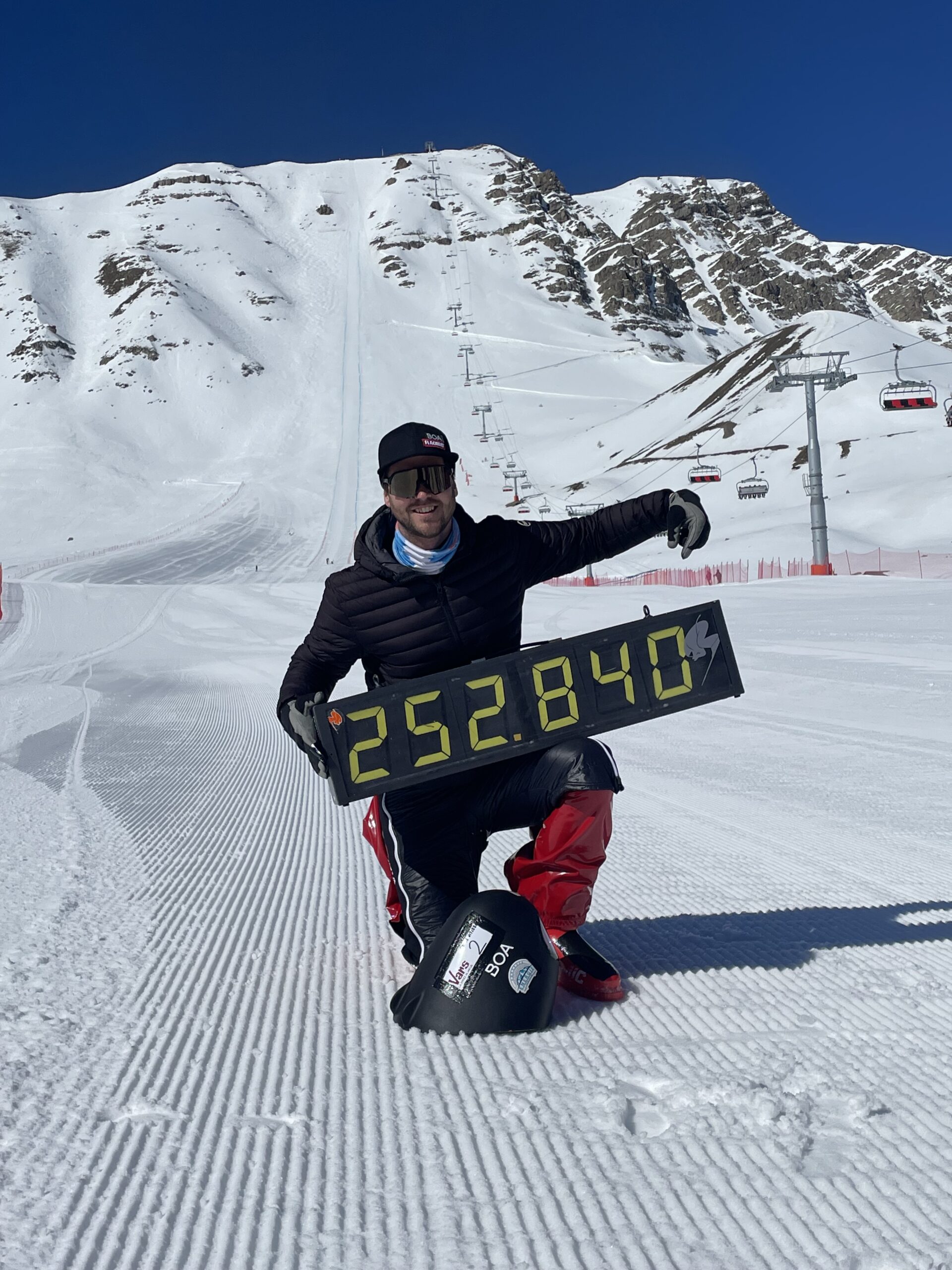 Person, die im Schnee kniet und ein Schild mit der Nummer 252.840 hält, mit verschneiten Bergen und Skiliften im Hintergrund.