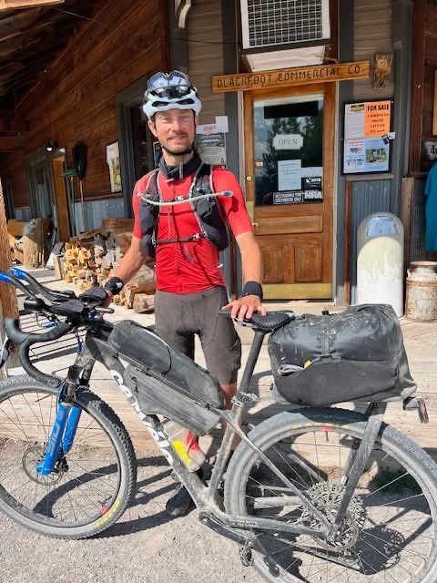 Ein Radfahrer in roter Kleidung steht mit einem beladenen Tourenrad vor einem rustikalen Gemischtwarenladen aus Holz.
