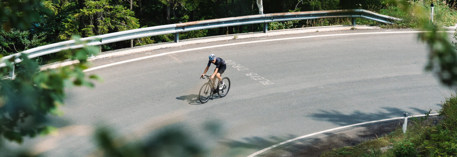 Ein Radfahrer mit Helm fährt allein auf einer kurvigen Bergstraße, umgeben von Bäumen und Grün.