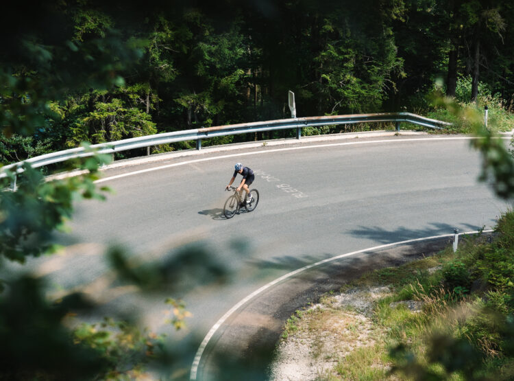 Ein Radfahrer mit Helm fährt allein auf einer kurvigen Bergstraße, umgeben von Bäumen und Grün.