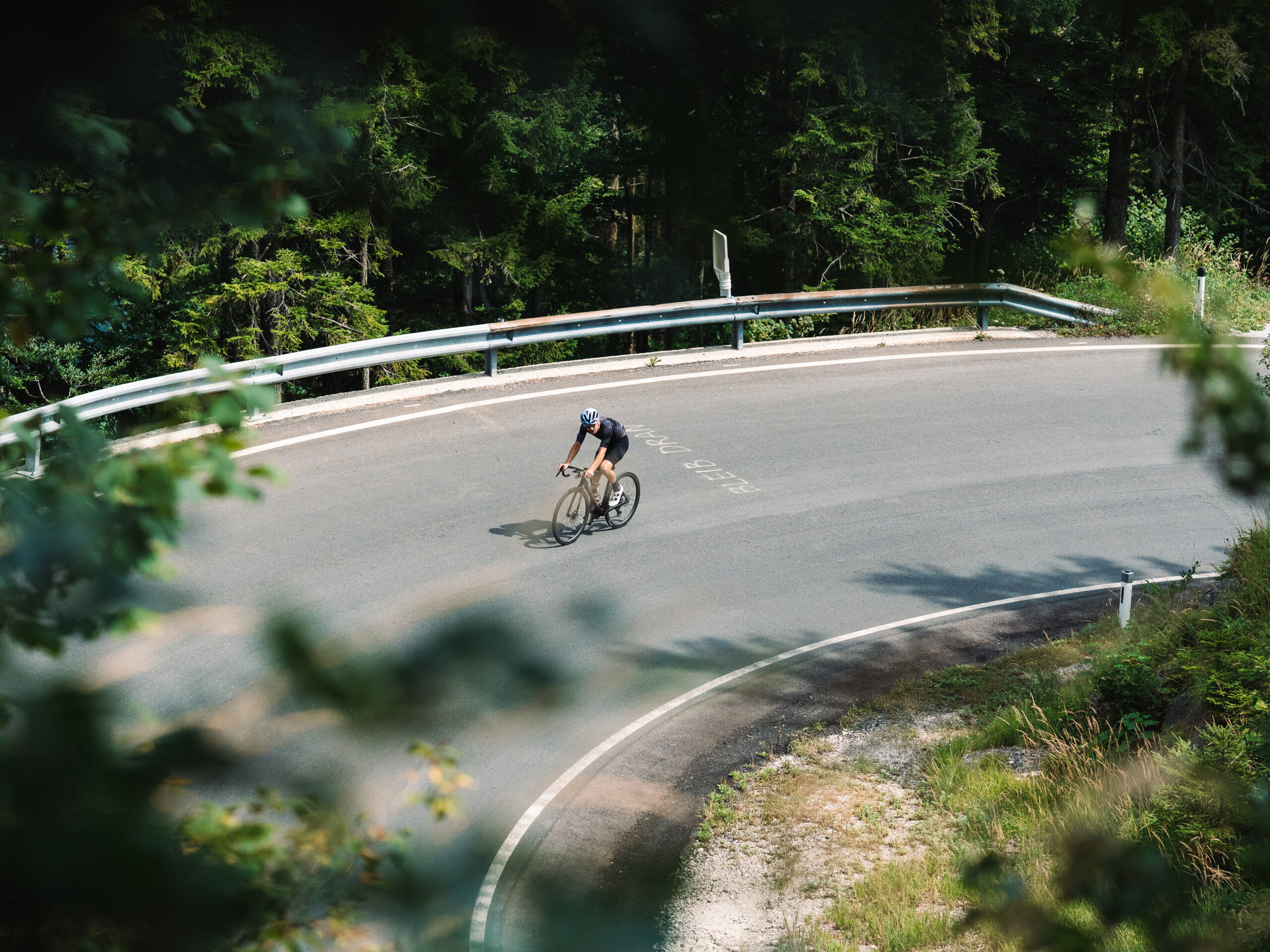 Ein Radfahrer mit Helm fährt allein auf einer kurvigen Bergstraße, umgeben von Bäumen und Grün.