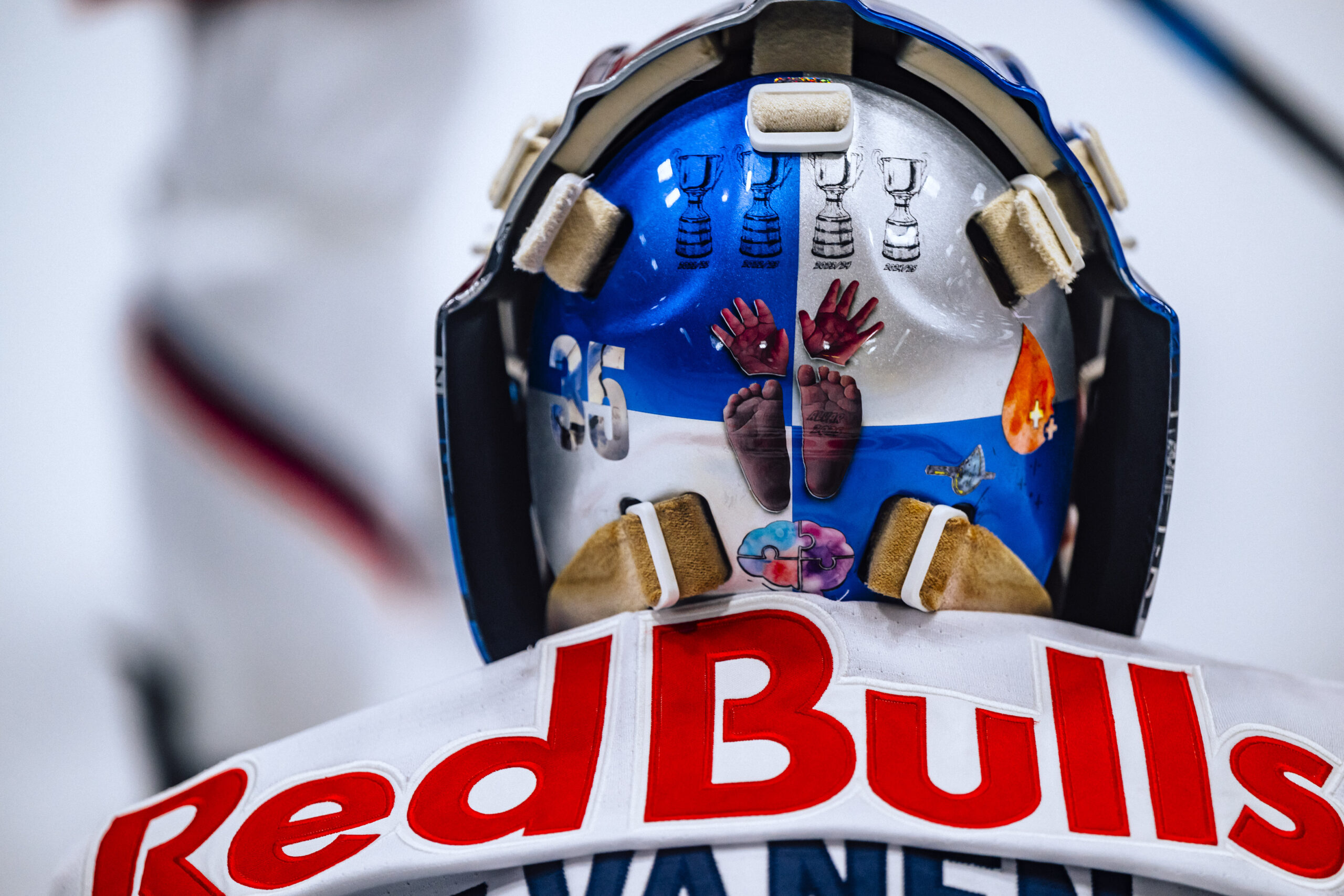 Ein Eishockeytorwart, der einen speziellen Helm mit Trophäen und Kunstwerken trägt, und ein Red Bulls-Trikot von hinten gesehen.