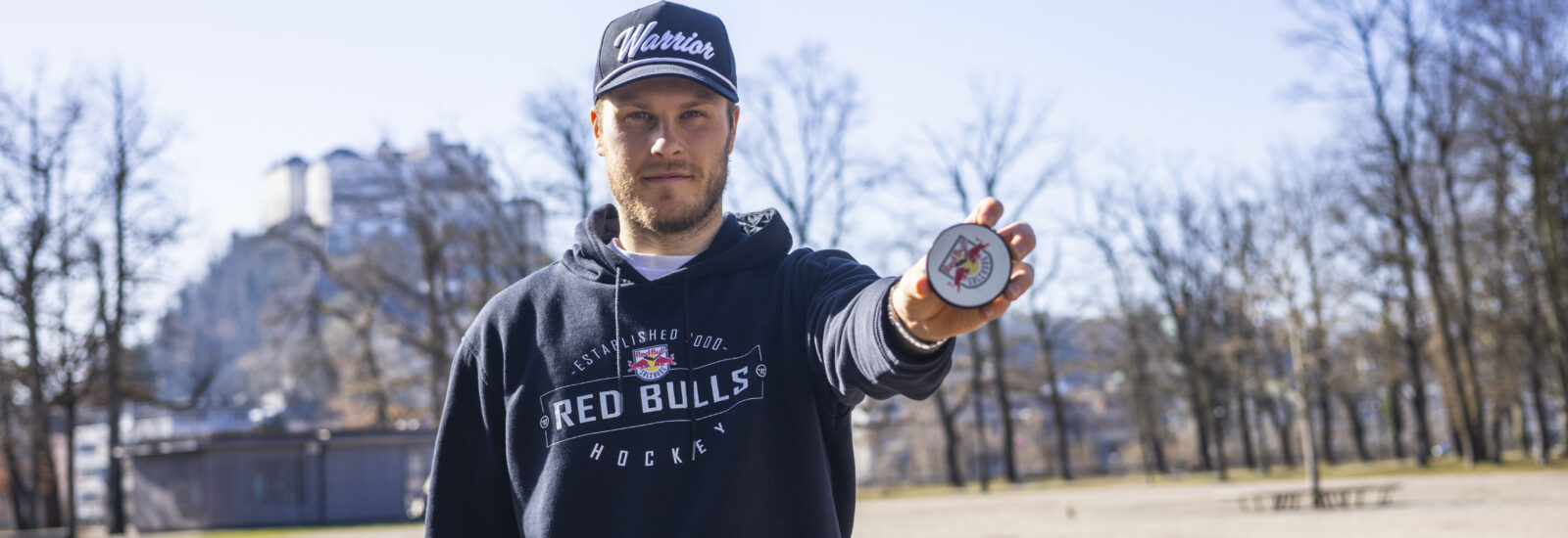 Ein Mann in einem Red Bulls Hockey-Kapuzenpulli hält in einem sonnigen Park eine Medaille vor sich her.