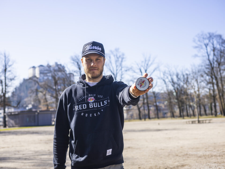 Ein Mann in einem Red Bulls Hockey-Kapuzenpulli hält in einem sonnigen Park eine Medaille vor sich her.
