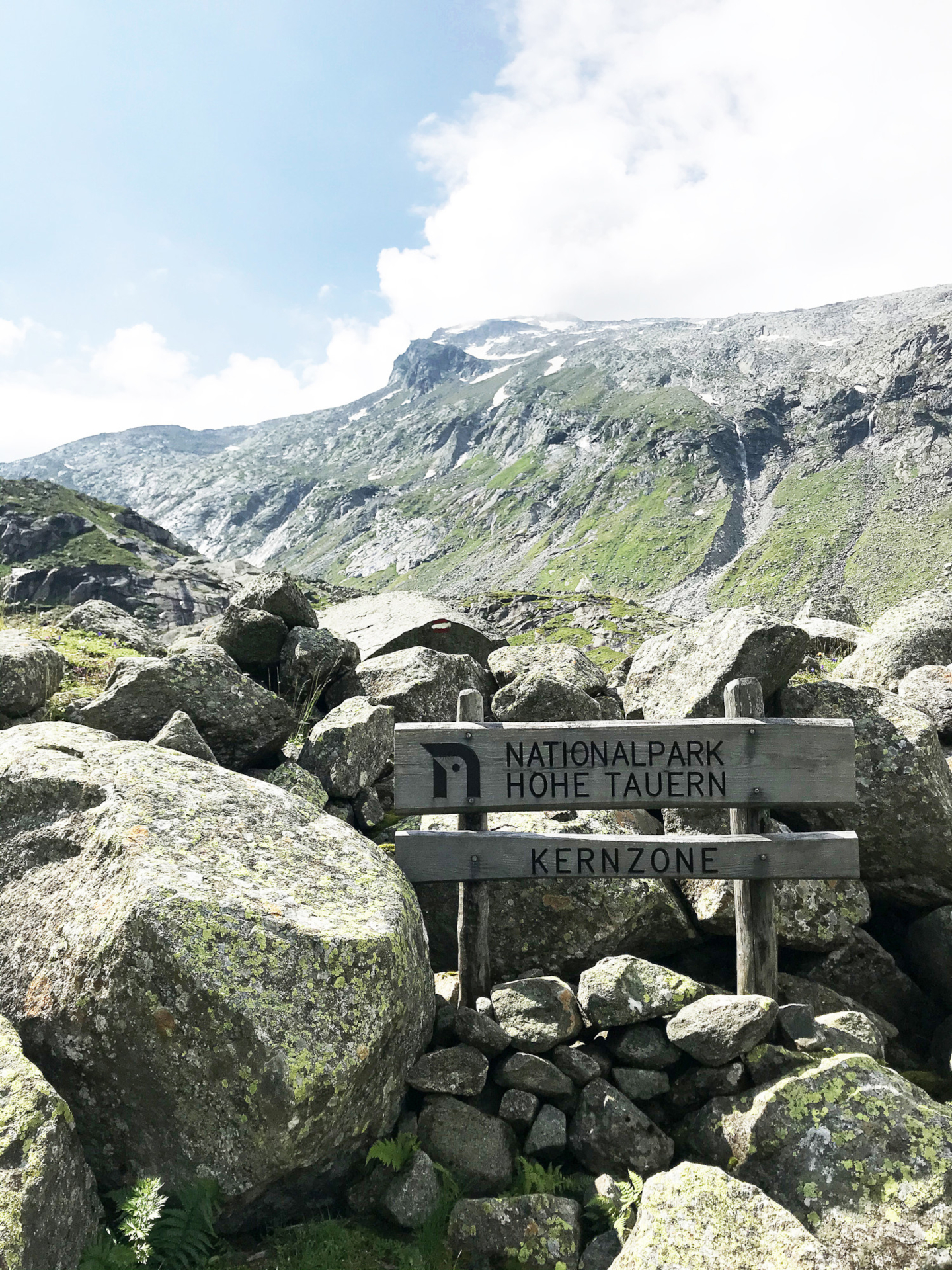 Holzschild mit der Aufschrift Nationalpark Hohe Tauern Kernzone zwischen Felsen, mit Bergen im Hintergrund.