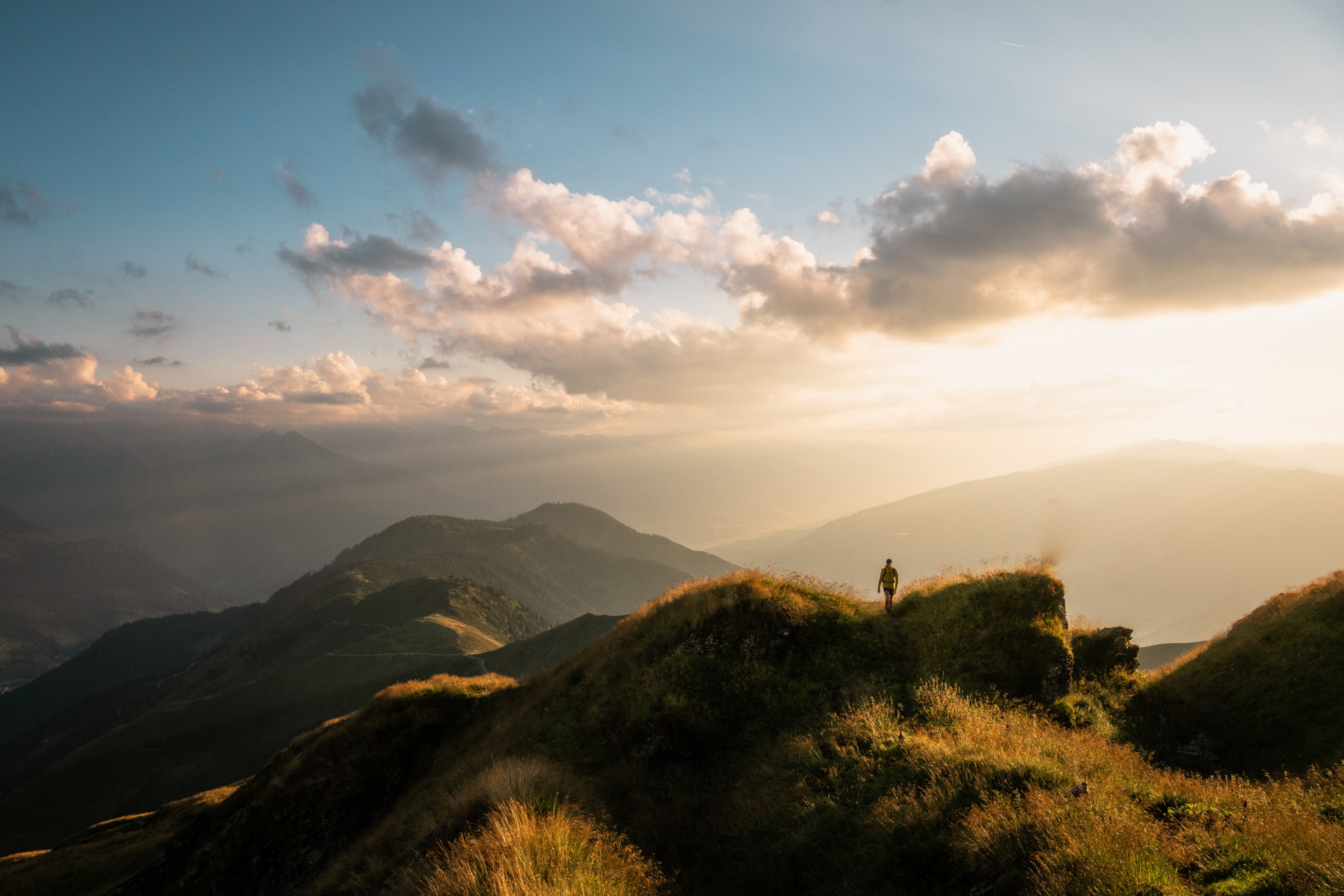 Person auf einem grasbewachsenen Bergrücken bei Sonnenuntergang mit Wolken und fernen Hügeln im Hintergrund.