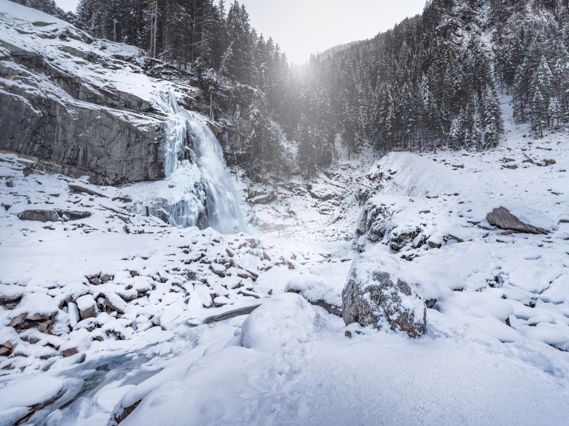 Ein gefrorener Wasserfall, umgeben von verschneiten Felsen und Kiefern an einem hellen Wintertag.