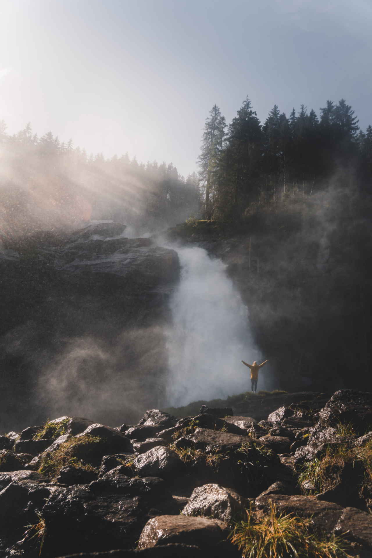 Eine Person steht mit erhobenen Armen in der N&auml;he eines nebligen Wasserfalls, umgeben von Felsen und hohen B&auml;umen im Sonnenlicht.