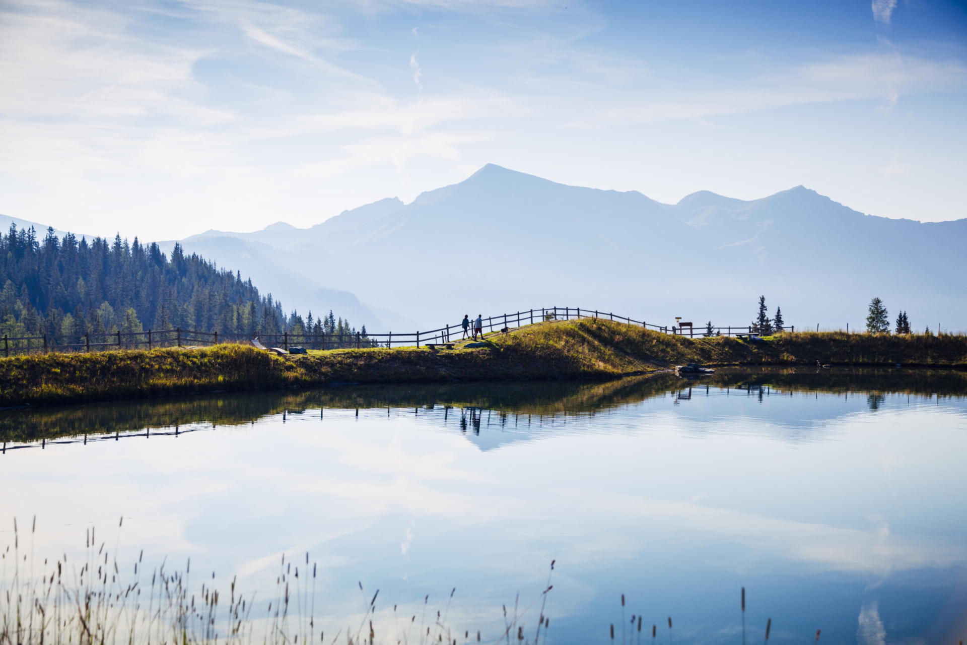 In einem ruhigen See spiegeln sich unter blauem Himmel Berge und ein Zaun, an dessen Ufer zwei Personen spazieren gehen.