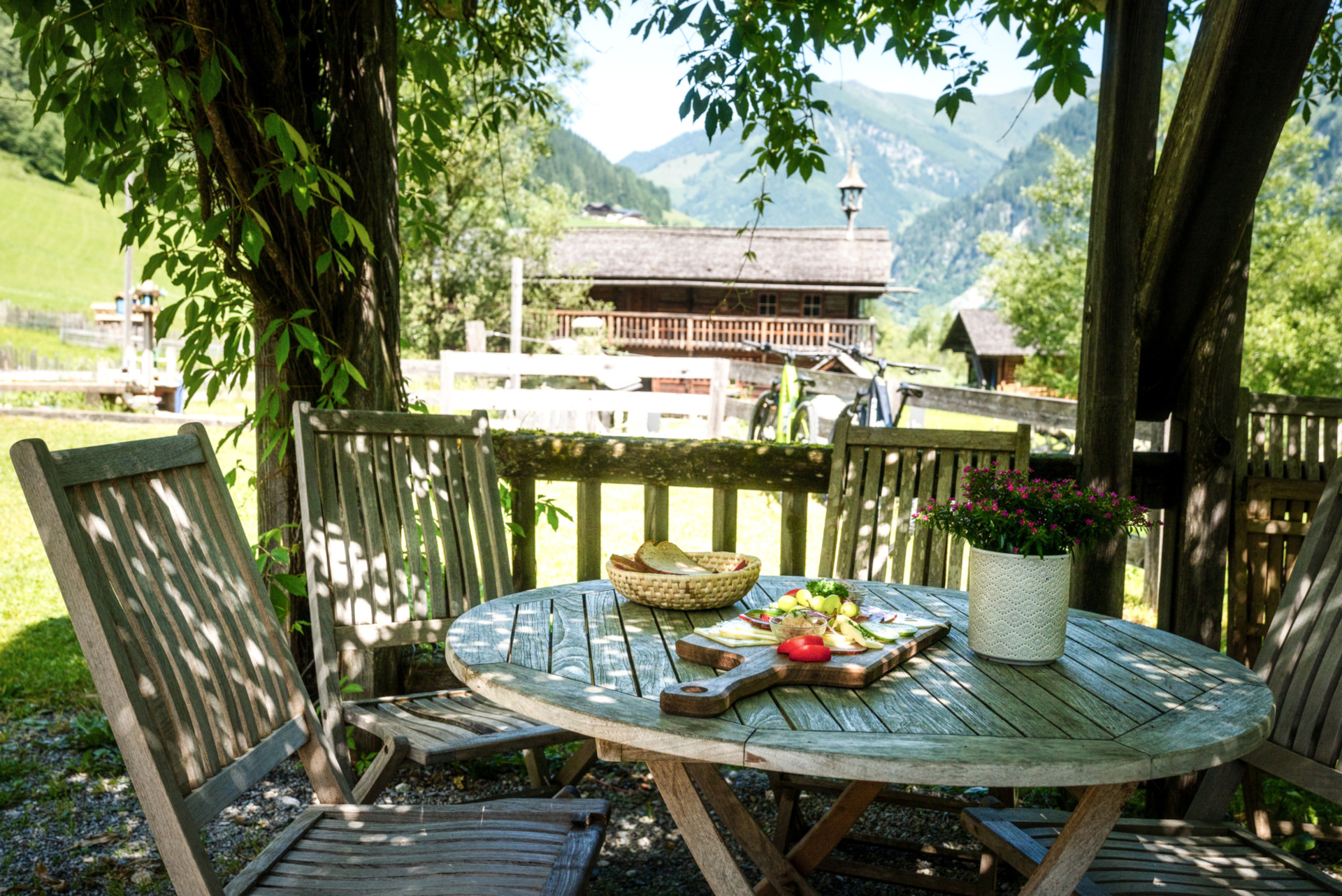 Holztisch mit Essen und Blumen auf einer Terrasse, mit Blick auf ein Bauernhaus und grüne Berge im Sommer.
