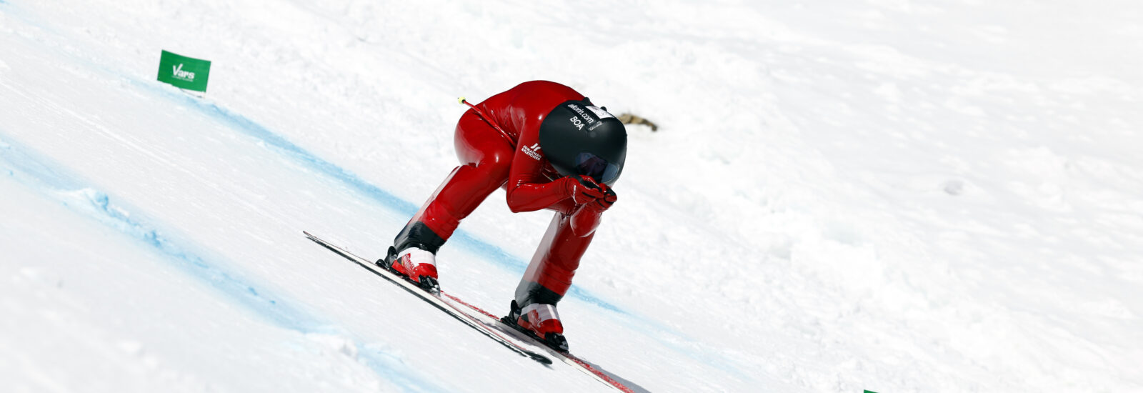 Ein Skifahrer im roten Anzug hockt tief, während er auf einer verschneiten Piste bergab fährt.