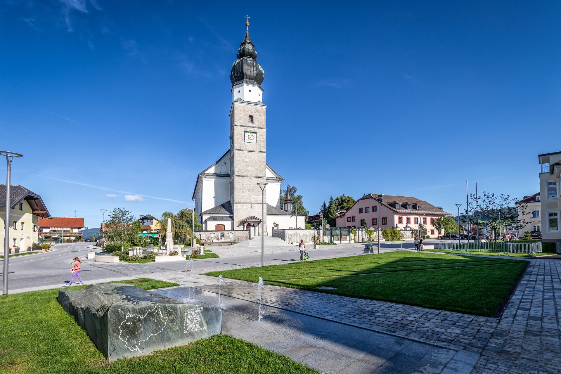 Ein Stadtplatz mit einer historischen Kirche, grünen Rasenflächen, einem Brunnen und Menschen, die an einem sonnigen Tag spazieren gehen.