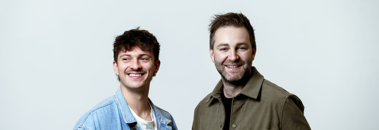 Zwei Männer stehen nebeneinander, lächelnd, vor einem schlichten weißen Hintergrund. Einer trägt eine Jeansjacke, der andere eine olivfarbene Jacke.
