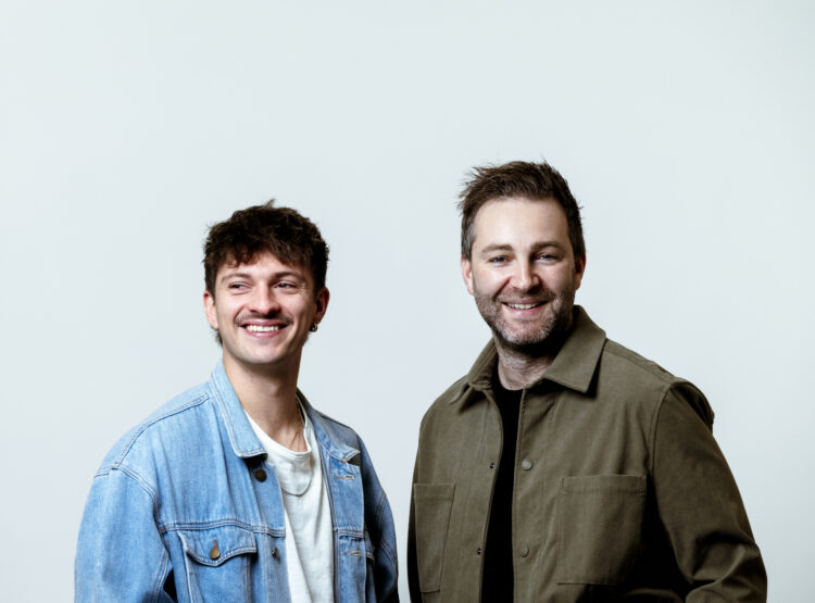 Zwei Männer stehen nebeneinander, lächelnd, vor einem schlichten weißen Hintergrund. Einer trägt eine Jeansjacke, der andere eine olivfarbene Jacke.