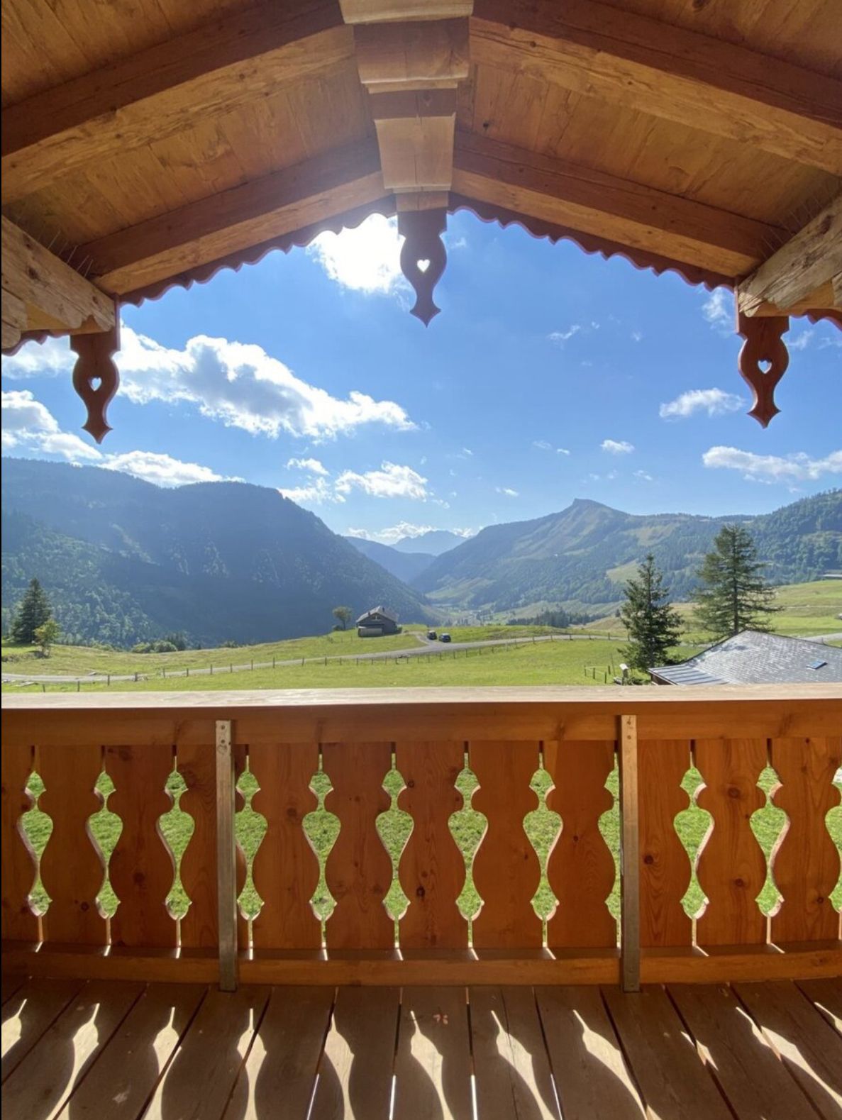 Blick von einem Holzbalkon auf gr&uuml;ne H&uuml;gel, B&auml;ume und ferne Berge unter blauem Himmel.